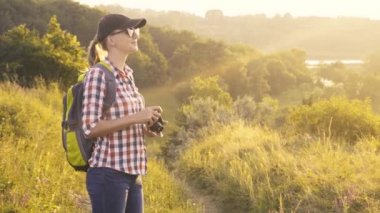 Sırt çantası ve fotoğraf makinesi olan turist kız resimli bir çayırda yürüyor ve manzaranın fotoğraflarını çekiyor..