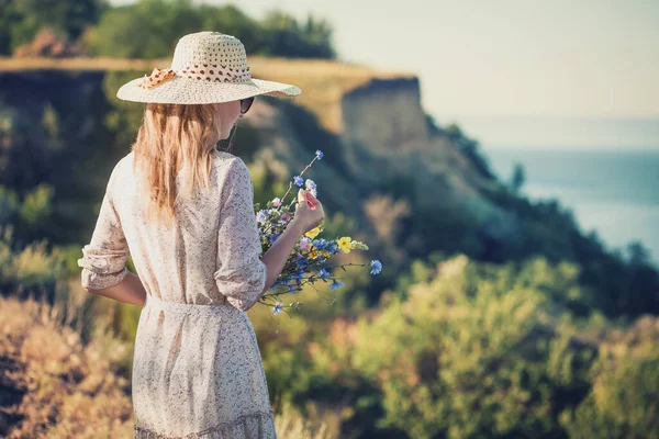 美しい若い女性が高い海の崖の上の帽子にレトロな帽子の花の花束 — ストック写真
