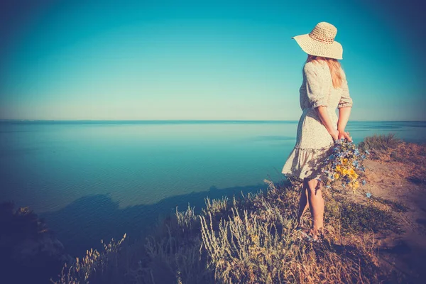 美しい若い女性が高い海の崖の上の帽子にレトロな帽子の花の花束 — ストック写真