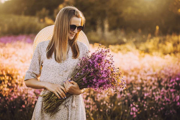 山の花の花束を背景に夕日と日当たりの良い開花草原に立っている若い女性 — ストック写真