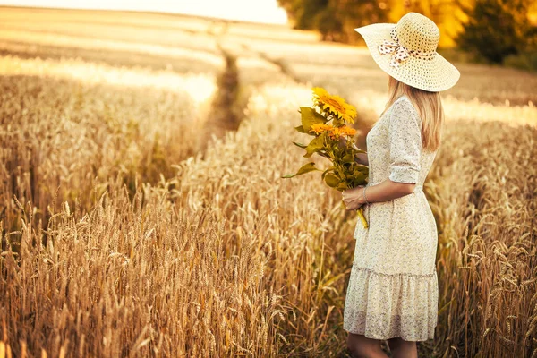 Žena se slunečnicemi v retro šatech a kloboukem v letním pšeničném poli — Stock fotografie