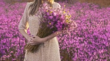 Bir buket kır çiçeğiyle güneşli bir çayırda duran genç bir kadın.