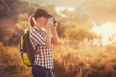 Sırt çantası ve kameralı turist kız resimli bir çayırda yürüyor ve manzaranın fotoğraflarını çekiyor. Turizm, açık hava aktiviteleri