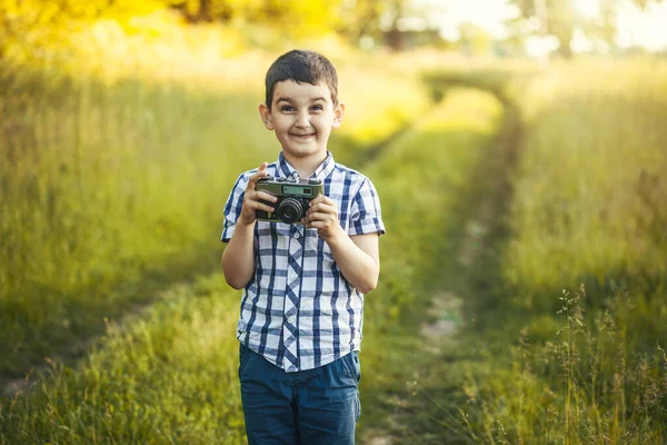 在阳光灿烂的草地上 一个带着复古的Dslr相机的漂亮的笑脸男孩 背景是夕阳西下 — 图库照片