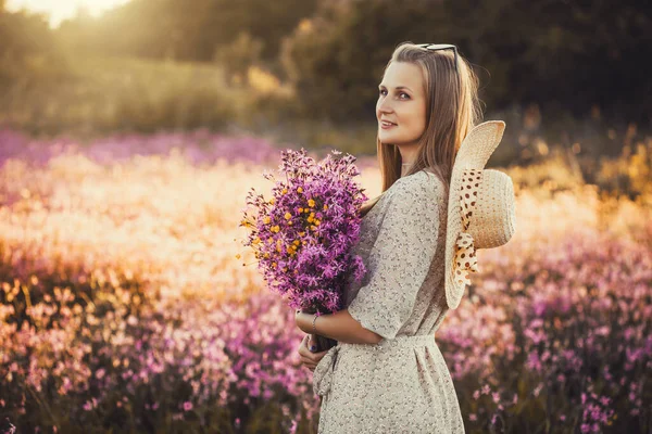 山の花の花束を背景に夕日と日当たりの良い開花草原に立っている若い女性 — ストック写真
