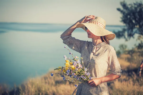 Mulher em um chapéu em um penhasco alto mar com um buquê de flores em um chapéu retro. — Fotografia de Stock