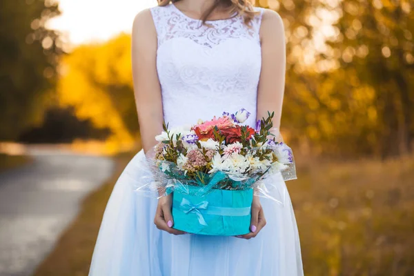 Mãos femininas com uma caixa de flores no fundo de um vestido de noiva branco. — Fotografia de Stock