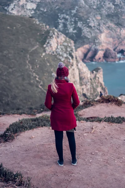 Mujer joven en un abrigo rojo contemplando el mar desde la cima de un acantilado —  Fotos de Stock