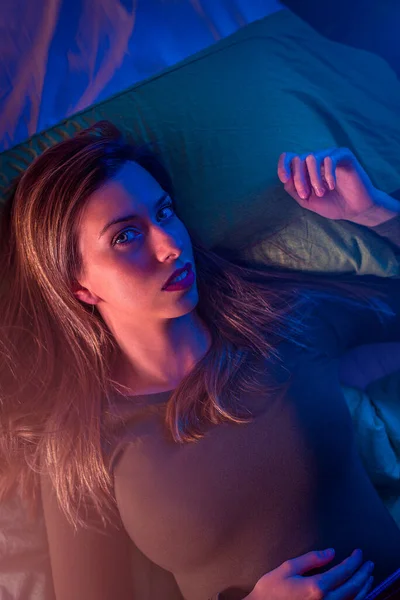 Portrait of a young woman on a bed from above with crossed colors from neon lights