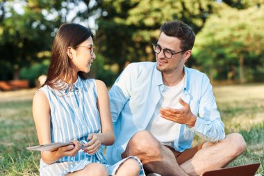 Dizüstü bilgisayarı ve tableti olan genç öğrenciler çimlerin üzerinde oturuyor. 