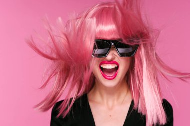 Pembe peruklu ve güneş gözlüklü genç bir kadın stüdyoda poz veriyor. 