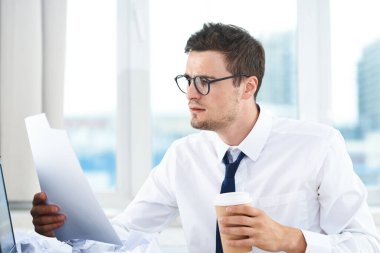 Yong yakışıklı işadamı elinde belge ve kahveyle ofiste oturuyor.