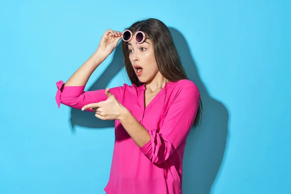 Young beautiful confused  woman in sunglasses pointing   in studio isolated on blue background