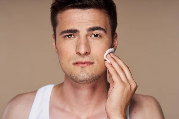 Studio shot. Young man cleaning his face with lotion  isolated on beige background