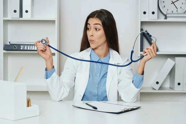 Young  confused woman doctor looking at stethoscope while   sitting at desk