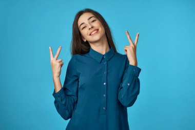 Stüdyoda zafer işaretleri olan genç bir kadın mavi arka planda izole edilmiş. 