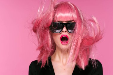 Pembe peruklu ve güneş gözlüklü genç bir kadın stüdyoda poz veriyor. 