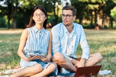 Dizüstü bilgisayarı ve tableti olan genç öğrenciler çimlerin üzerinde oturuyorlar. 