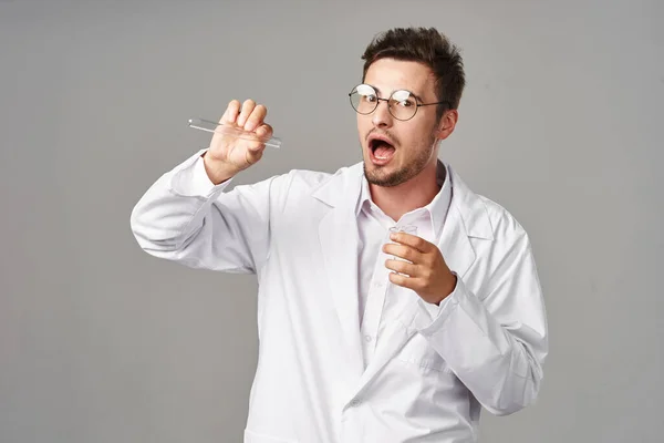 Young handsome  confused doctor in lab coat with test tubes on grey background