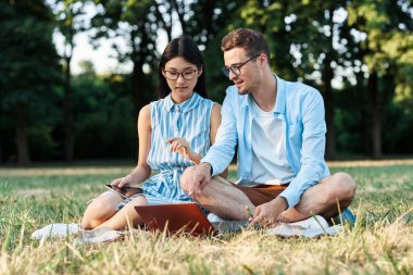 Dizüstü bilgisayarı ve tableti olan genç öğrenciler çimlerin üzerinde oturuyorlar. 