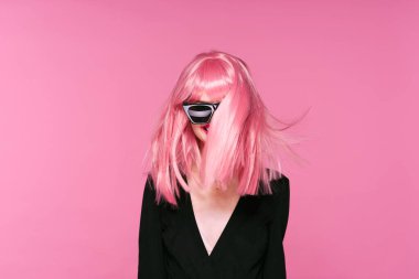 Pembe peruklu ve güneş gözlüklü genç bir kadın stüdyoda poz veriyor. 