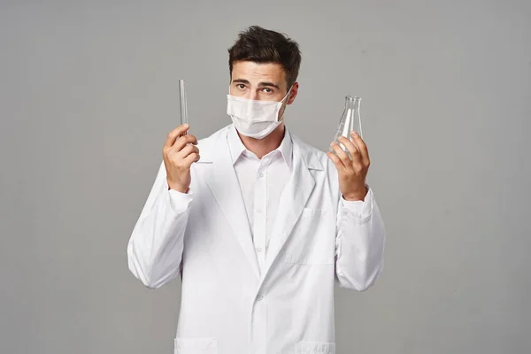 Young handsome doctor in face mask holding  test tubes on grey background
