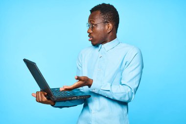 Genç Afrikalı sürpriz iş adamı dizüstü bilgisayarını mavi arka planda tutuyor.