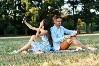 Dizüstü bilgisayarı ve tableti olan genç öğrenciler çimlerin üzerinde oturuyorlar. 