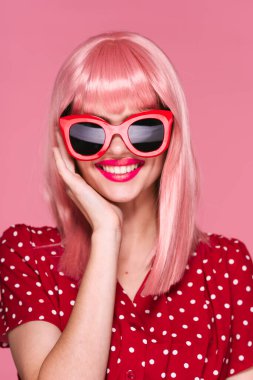 Pembe peruklu ve güneş gözlüklü genç bir kadın stüdyoda poz veriyor. 