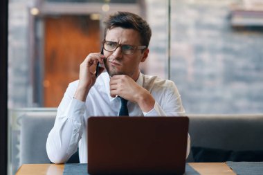 Genç yakışıklı iş adamı ofiste dizüstü bilgisayarla çalışıyor ve akıllı telefondan konuşuyor. 