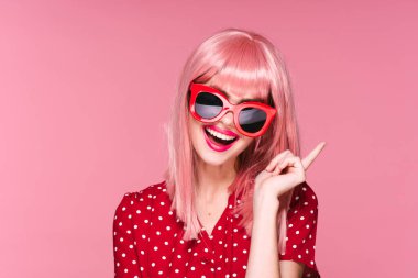 Pembe peruklu ve güneş gözlüklü genç bir kadın stüdyoda poz veriyor. 