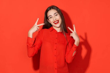 Stüdyo çekimi. Kırmızılı genç ve güzel bir kadın. Kırmızı arka planda izole edilmiş zafer işaretleriyle.