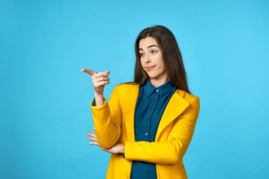 Sarı ceketli genç ve güzel bir kadın mavi arkaplan ile stüdyoyu işaret ediyor..