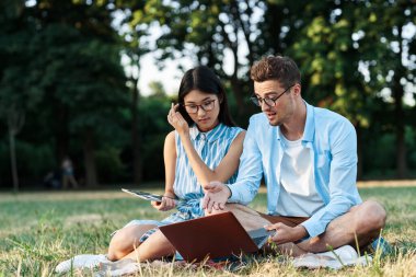Dizüstü bilgisayarı ve tableti olan genç öğrenciler çimlerin üzerinde oturuyorlar. 