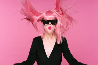 Pembe peruklu ve güneş gözlüklü genç bir kadın stüdyoda poz veriyor. 