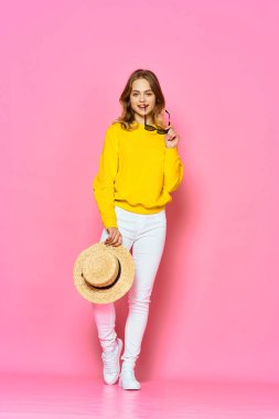Stüdyoda pembe arka planda şapka ve güneş gözlüğü tutan genç ve güzel bir kadın.      