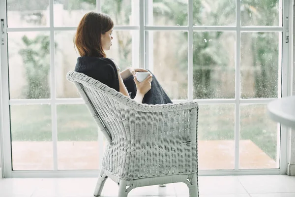 Kobieta na krześle w pobliżu okna w kratkę na ramionach filiżankę kawy — Zdjęcie stockowe