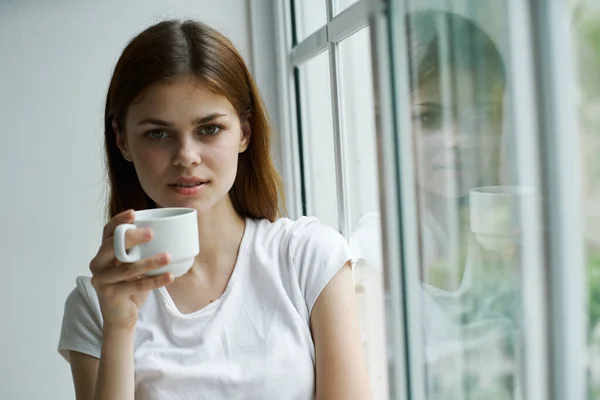 Спокойная женщина с чашкой возле окна смотрит в камеру — стоковое фото