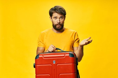 Sarı tişörtlü bir adam elinde kırmızı bir bavul tutuyor.