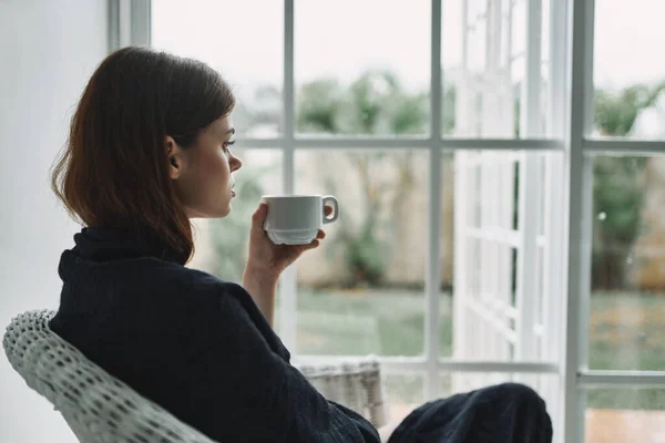 Mujer en una silla con una taza de café mirando por la ventana —  Fotos de Stock