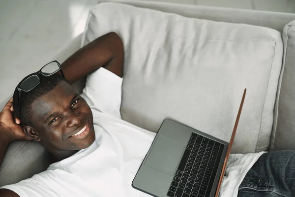 Um afro-americano com um laptop está no sofá e óculos — Fotografia de Stock