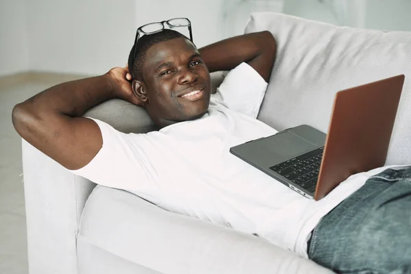 A man lies on the couch in front of a laptop African appearance leisure Stock Picture