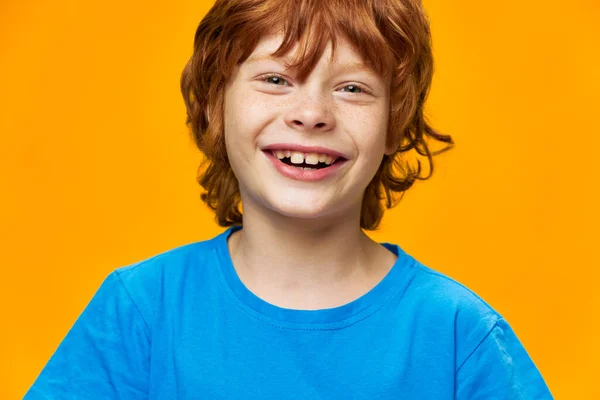 A funny redhead boy smiles into the camera close-up blue t-shirt yellow background — Stock Photo, Image