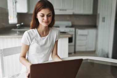 Mutfakta dizüstü bilgisayarın önündeki masada oturan bir kadın.