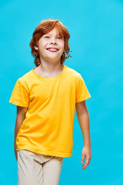 Happy red hair child in a T-shirt and trousers on a blue background shows his tongue — Stock Photo, Image