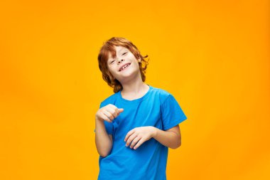  Mavi tişörtlü kızıl saçlı çocuk elleriyle gülümsüyor ve jestler yapıyor.