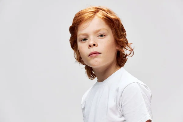 Söt rödhårig pojke i vit t-shirt ser rakt på grå — Stockfoto