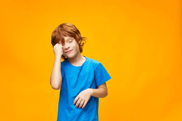 Rödhårig pojke blå t-shirt gul bakgrund fräknar och röra ansiktet med handen ett vackert leende. — Stockfoto