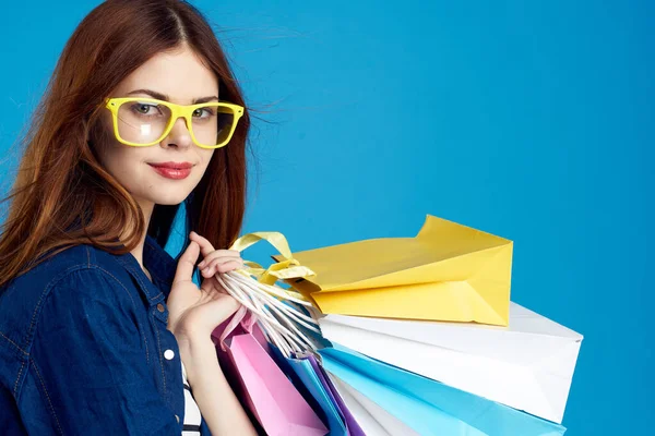 Mujer de moda es ir de compras con paquetes en un fondo azul en una camiseta a rayas y chaqueta, gafas en la cara —  Fotos de Stock