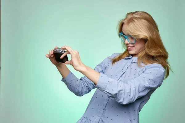 Mulher fotógrafa loira em óculos com uma câmera em suas mãos em um fundo verde em uma camisa azul — Fotografia de Stock